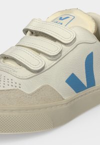 Gros plan d'une basket blanche avec trois bandes Velcro, des détails en daim beige et un grand logo bleu en forme de V sur le côté.