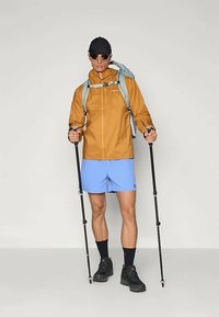Brown waterproof jacket, light blue shorts, black shoes. Holding trekking poles, wearing sunglasses and a cap. Backpack with straps.