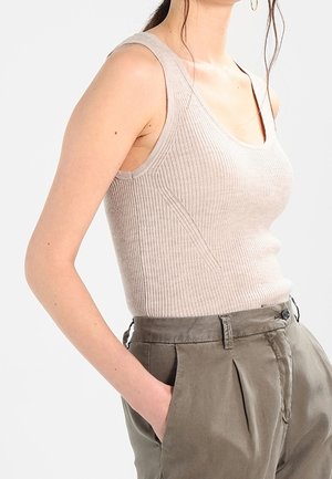 Vrouw met een beige geribde mouwloze top en hooggetailleerde olijfgroene broek met één hand in de zak, tegen een effen achtergrond.