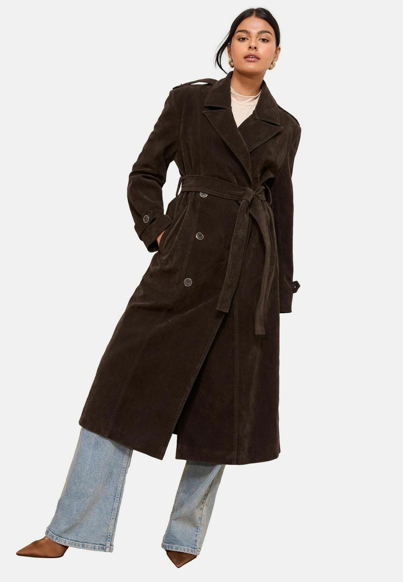 Femme portant un trench-coat ceinturé marron foncé sur un jean bleu clair et des chaussures marron pointues, debout avec une main dans la poche.