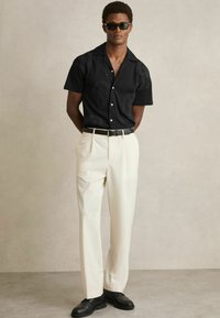Cămășă neagră cu mâneci scurte și model texturat, cu butoane albe, purtată cu pantaloni crem și pantofi negri. Modelul poartă ochelari de soare închiși la culoare.