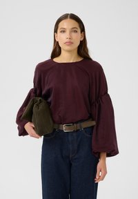 Burgunderfarget satengbluse med voluminøse pufferm, sammen med høyden på mørk denimjeans og et brunt belte, holder en grønn veske.