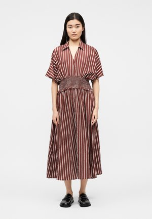 Fiatal nő áll barna és fehér, függőlegesen csíkos ruhában, melynek összegyűrt dereka van, fekete beszállócipőben, fehér háttér előtt.