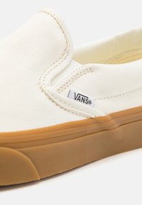 Bijela platnena slip-on cipela s gumenim potplatom i Vans logotip oznakom sa strane blizu elastične gumice.
