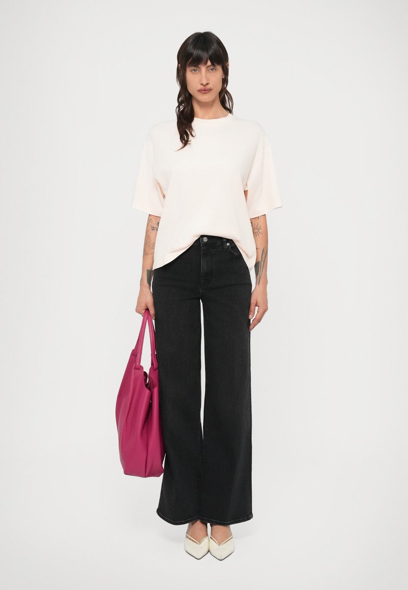 Czarne dżinsy o szerokich nogawkach w połączeniu z jasnoróżowym, oversize'owym t-shirtem. Trzyma teksturowaną różową torbę i nosi białe buty z ostrymi noskami.