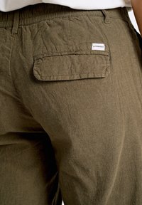 Pantalones de lino verde oliva con una textura arrugada, que cuentan con un bolsillo trasero y una pequeña etiqueta de marca. Cintura elástica para un ajuste y comodidad.