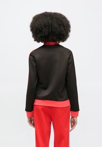 Must spordijakk punaste detailidega ja soonikkoes äärega, disainitud pikkade varrukate ja tõstetava kraega. Sile tekstuur, minimaalne viimistlus.