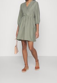 Abito avvolgente verde oliva con maniche a sbuffo e vita cinchata. Abbinato a sandali espadrilles beige e a una borsa rosa chiaro.