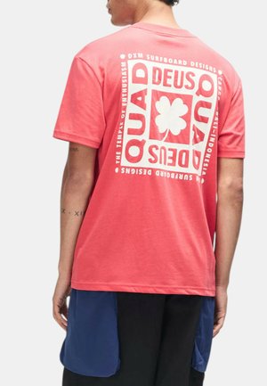 Persona con una camiseta rosa coral con gráfico de un cuadrado blanco y texto en la espalda, combinada con pantalones cortos negros y azules.