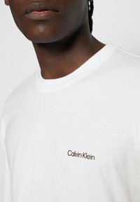Nahaufnahme einer Person, die ein weißes T-Shirt mit dem Aufdruck "Calvin Klein" auf der Brust trägt.