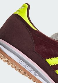Burgundi színű velúrmárkájú sneaker, neon sárga részletekkel, melynek strukturált felülete van, rózsaszín középtalpa és zig-zag mintázatú gumitalpa van.