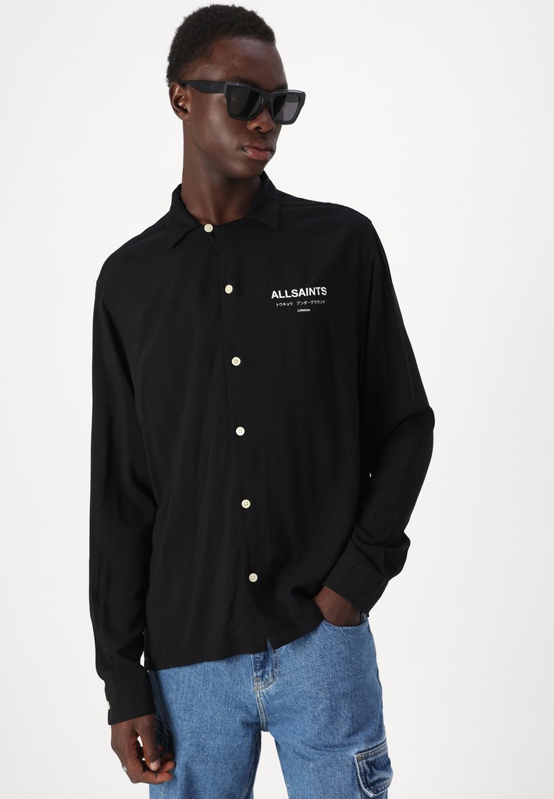 Zwarte button-up shirt met een relaxte pasvorm, voorzien van een logo aan de voorkant, kleine witte knopen en lange mouwen. Draagt met een spijkerbroek.