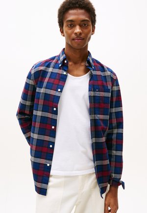 Ung man som bär en blå, röd och vit rutig skjorta över en vit linne och vita byxor, stående mot en enkel ljus bakgrund.