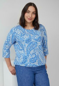 Blau und weißes Oberteil mit Paisleymuster, rundem Ausschnitt und drei Viertelärmeln. Kombiniert mit blauen Jeans.