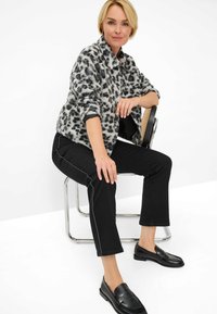 Faux fur jacket met een grijs en zwarte luipaardprint, zwarte slim-fit broek met witte bies, en zwarte loafers, zittend op een stoel.