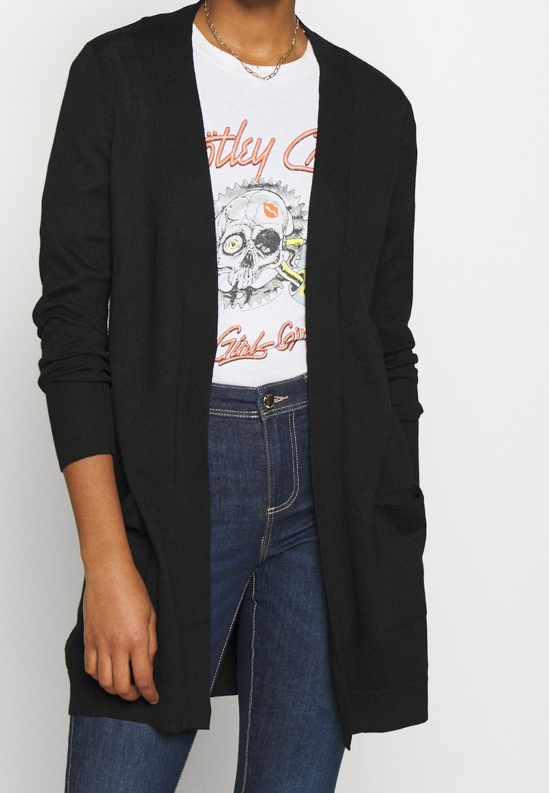 Personne portant un jean bleu foncé, un t-shirt blanc avec un motif de crâne et un cardigan noir ouvert sur le devant.
