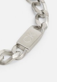 Silver metal chain bracelet with a rectangular clasp featuring engraved "CK." Links are interwoven with a polished finish and textured accents.