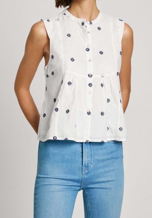 Kobieta ubrana w bezrękawnikową białą bluzkę z granatowymi haftowanymi kropkami i jasnoniebieskie dżinsy z wysokim stanem, stojąca na neutralnym tle.