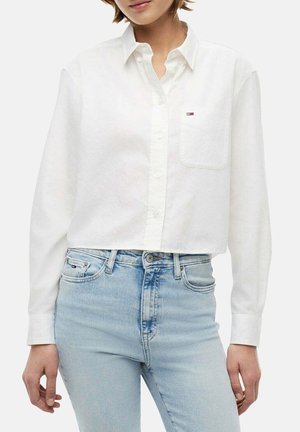 Donna che indossa una camicia bianca corta con bottoni e taschino sul petto, e jeans azzurri a vita alta, in piedi davanti a uno sfondo uniforme.