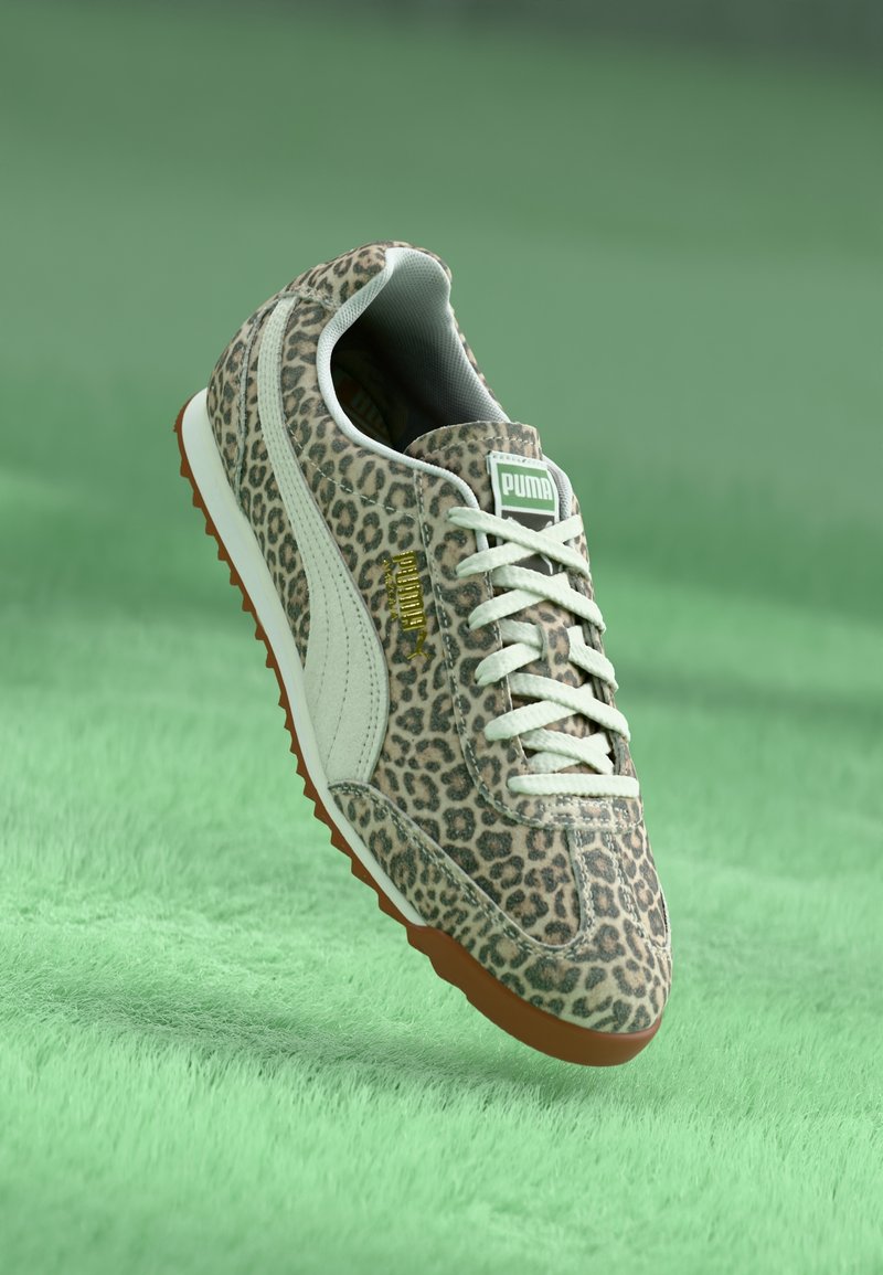 Baskets Puma imprimé léopard avec des rayures blanches, des lacets blancs et une semelle gomme, montrées flottant au-dessus d'une surface verte douce.