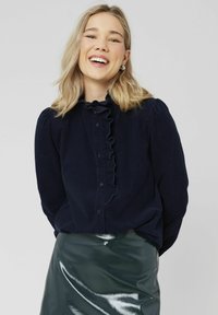 Chemise boutonnée bleu marine avec détail de col volanté, présentant un tissu texturé et des manches bouffantes. Associée à une jupe en cuir vert brillant.