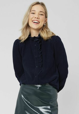 Chemise boutonnée bleu marine avec détail de col volanté, présentant un tissu texturé et des manches bouffantes. Associée à une jupe en cuir vert brillant.