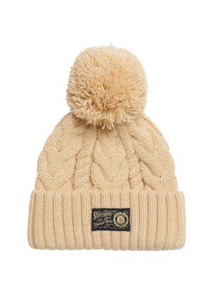 Gorro de punto beige con un patrón de trenzas texturizadas, que presenta un gran pompón en la parte superior y una etiqueta negra con detalles dorados en el puño.