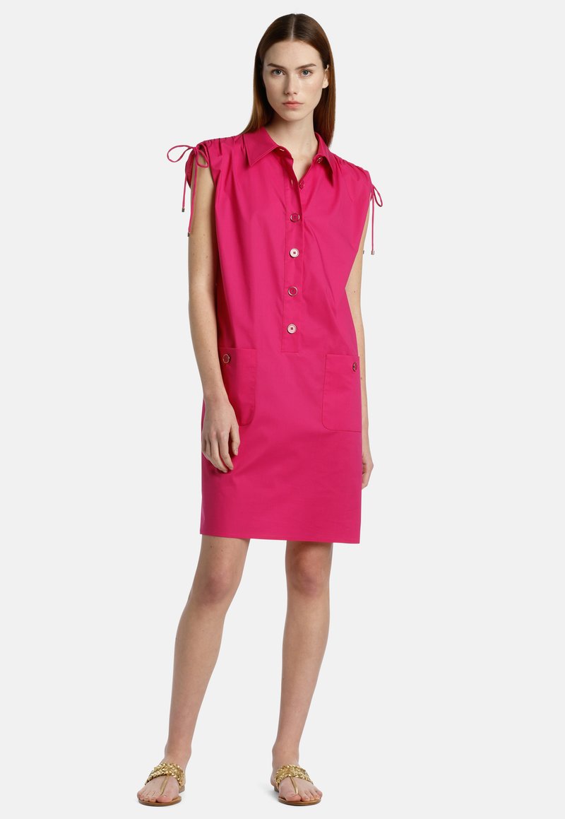Abito di cotone rosa con bottoni davanti, spalline e due tasche frontali. Tessuto morbido e design fino al ginocchio. Indossato con sandali flat.
