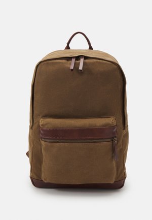 Sac à dos en toile marron avec poche frontale zippée et détails en cuir, présenté debout sur un fond blanc.