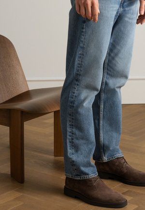 Lichtblauwe denim jeans met een relaxte pasvorm, gecombineerd met bruine suède laarzen. Op de achtergrond is een houten stoel te zien op een houten vloer.