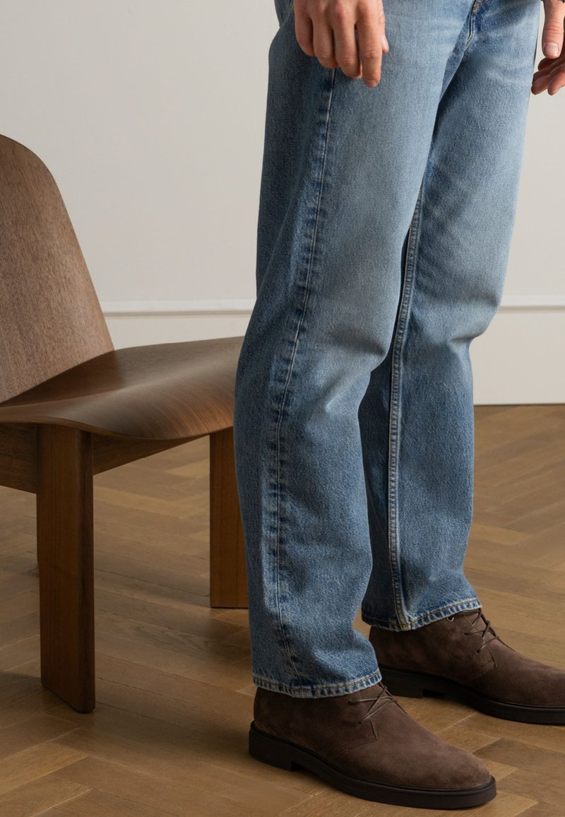Lichtblauwe denim jeans met een relaxte pasvorm, gecombineerd met bruine suède laarzen. Op de achtergrond is een houten stoel te zien op een houten vloer.