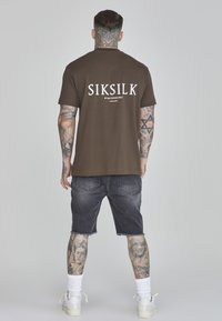 Camiseta de algodón marrón con texto blanco en la parte posterior. Combinada con pantalones cortos de mezclilla oscura, que revelan tatuajes detallados en las piernas, y zapatillas blancas.