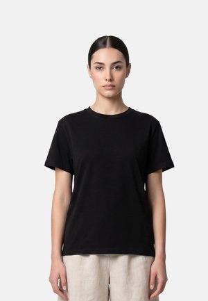 Junge Frau mit dunklen, zurückgebundenen Haaren, die ein schlichtes schwarzes Kurzarm-T-Shirt und hellfarbene Hosen trägt, steht vor einem weißen Hintergrund.