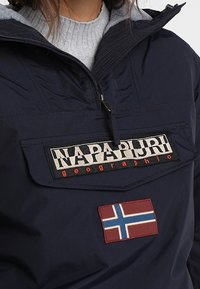 Veste isolante bleu marine avec une poche frontale, présentant un grand patch logo et un emblème de drapeau rouge avec une croix bleue. Tissu texturé.