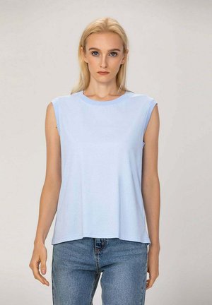 Femme blonde portant un haut sans manches bleu clair et un jean bleu, debout et faisant face à l'avant sur un fond uni.