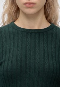 Gros plan d'une personne portant un pull torsadé vert foncé, visible du cou jusqu'à la partie supérieure de la poitrine et une partie des cheveux blonds clairs ondulés.