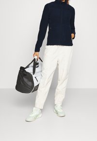 Giacca in fleece navy con zip e colletto alto, pantaloni tapered beige chiaro e scarpe da ginnastica multicolori. Porta una borsa a tracolla nera e bianca.