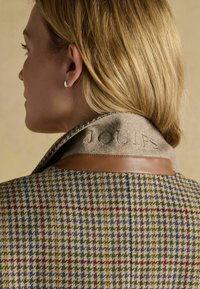 Vlnitá blendová bunda s houndstooth vzorom v zelenej, modrej a červenej farbe. Obsahuje krémový plsťový golier s vyrazeným nápisom "JOULES" a hnedé kožené akcenty.