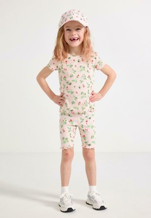 Smilende pige med hænderne på hofterne iført lyserød blomsterkasket, matchende skjorte og shorts, hvide sokker og sneakers på en hvid baggrund.