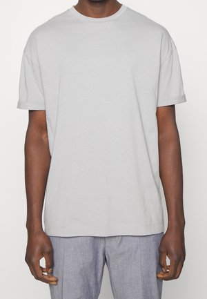 Mężczyzna ubrany w prostą jasnoszarą koszulkę z krótkim rękawem i jasnoszare spodnie, stojący z ramionami luźno opuszczonymi wzdłuż ciała na neutralnym tle.