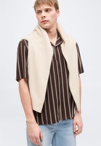 Νέος άνδρας που φοράει καφέ κοντομάνικη ριγέ μπλούζα, ανοιχτό μπεζ πουλόβερ χωρίς μανίκια περασμένο στους ώμους, και ανοιχτό μπλε τζιν, κοιτάζοντας προς τα αριστερά.