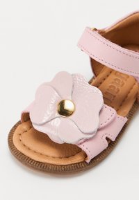 Rosa lädersandal med en stor, glänsande rosa blommor embellering med en gyllene mitt. Sulan är brun med texturerade detaljer.