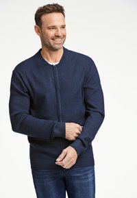 Marinblå stickad zip-up cardigan med ribbade muddar och nederkant, med rund halsringning och slät textur. Bärs över en vit skjorta.