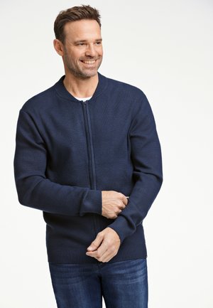 Cardigan in maglina blu navy con zip, maniche a costine e orlo a costine, caratterizzato da un colletto rotondo e una trama liscia. Indossato sopra una camicia bianca.