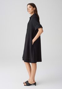 Vestito camicia nero a coste con maniche corte, tasche laterali e una vestibilità comoda. Abbinato a mule nere con tacco aperto.