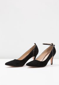 Paire d’escarpins en daim noir à bout pointu, avec bride à la cheville et doublure intérieure beige sur surface blanche et fond gris clair.