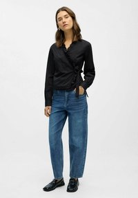 Frau in einer schwarzen, langärmligen Wickelbluse mit Rüschen, blauen hoch taillierten Jeans und schwarzen Slippern, die mit einer Hand in der Tasche steht.