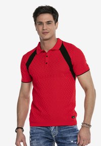 Rotes Poloshirt mit einem strukturierten Zickzackmuster, schwarzen Seitenelementen und drei Knöpfen am Kragen; kombiniert mit ausgeblichenen blauen Jeans.