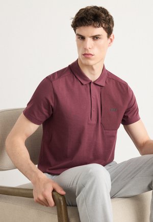 Burgundi polo särk, millel on nööbiga kaelus ja väike rinna tasku, valmistatud siledast kangast, kantud heledate hallide pükstega.