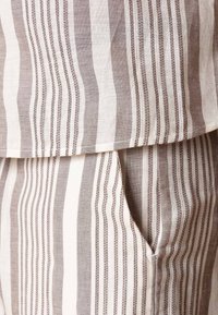 Tissu rayé avec des lignes beige clair et foncé alternées. Présente une couture texturée le long des bords, créant un aspect structuré.